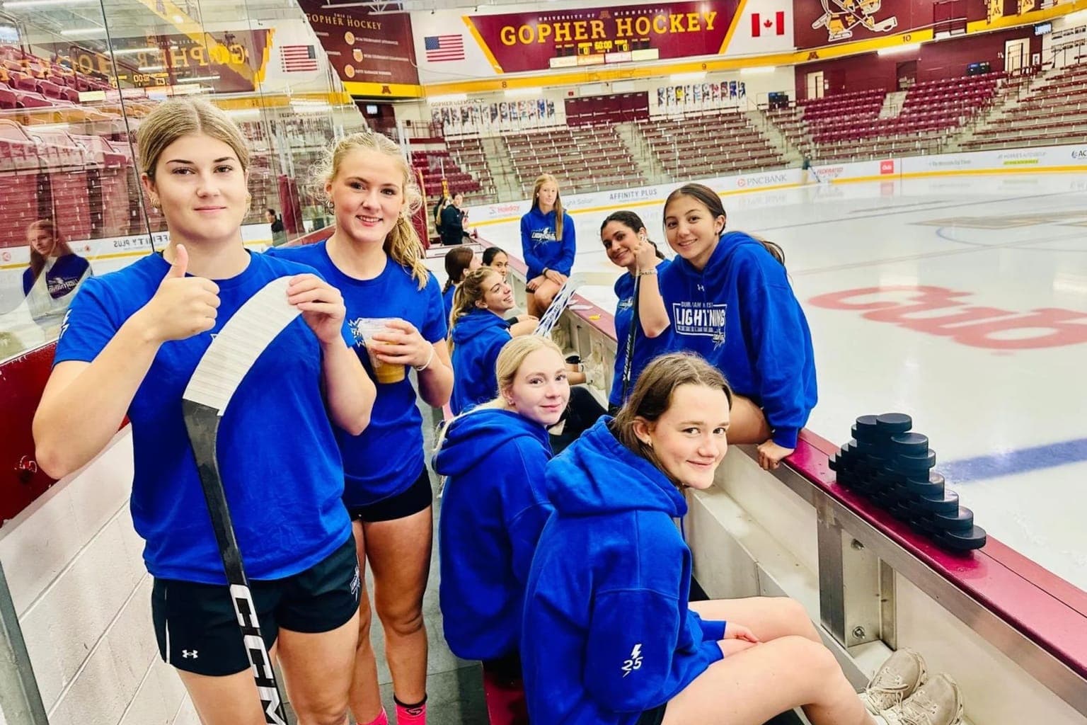 Durham West Junior Lightning players in blue team hoodies and t-shirts smile from the bench at the University of Minnesota's Ridder Arena during the NCAA Division I Trip.